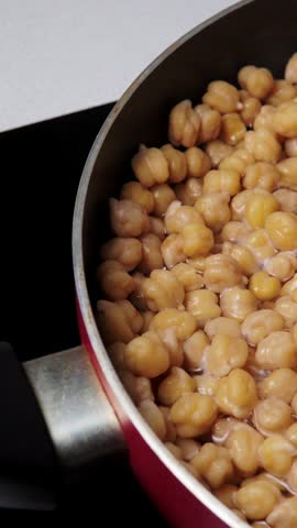 Vertical video close up view of round garbanzo beans boiling with bubbles in a pot on stove. Ideal for hummus preparation, vegan cooking
