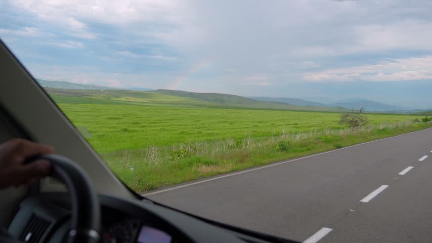 Beautiful view from a car driving on an asphalt road through a scenic countryside with green fields, rolling hills, and a faint rainbow appearing in the sky after the rain