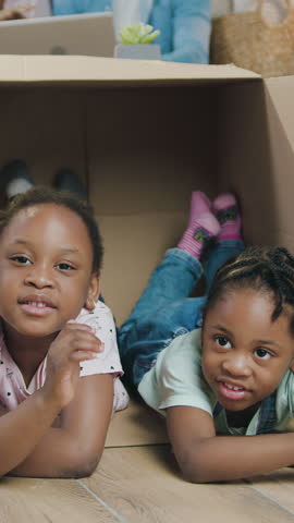 Lovely happy joyful little two african american sisters having fun together while climbing inside into cardboard packing after relocation with parents in their new house