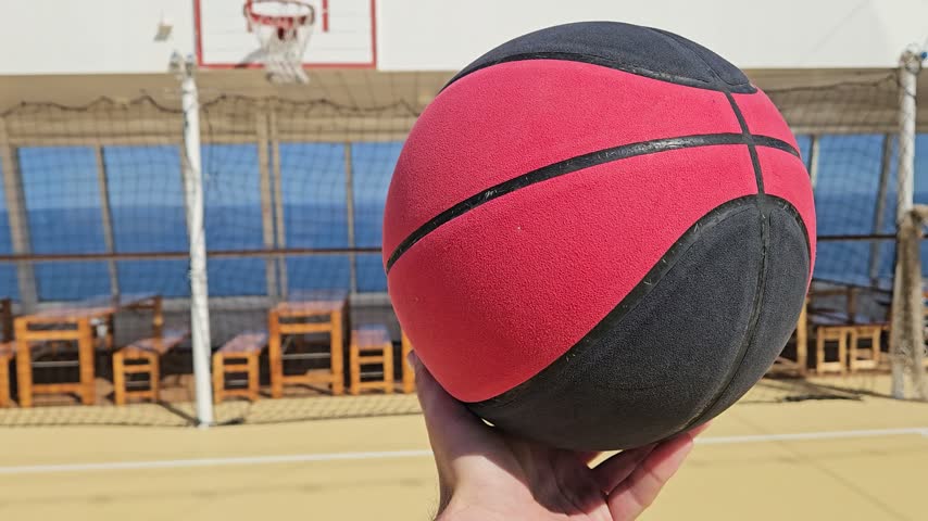 Hand holds red and black basketball. Basketball court with hoop and netting visible. Clear blue sky behind the cruise ship