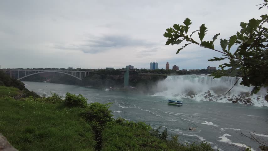 The Rainbow Bridge and the mist of the American Falls across the Niagara gorge, with a tour boat on the river, the Horseshoe Falls entering at the right and oak branches in the foreground