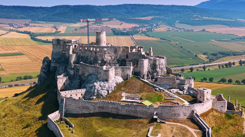 Hilltop castle under construction. A large castle on a hill with construction activity nearby. The landscape includes fields and hills in the background.