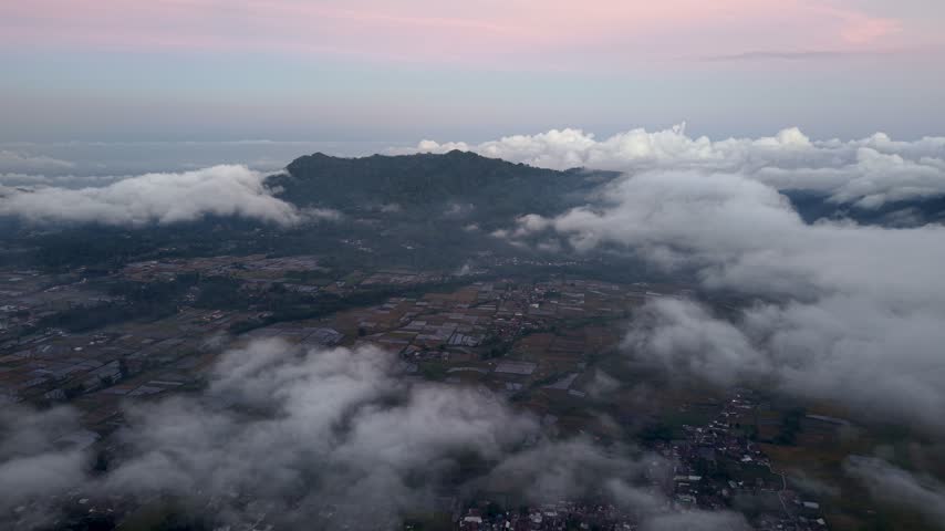 Misty sunrise over Pronojiwo highlands with clouds drifting above rice paddies and scattered village lights.