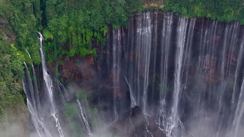 Powerful waterfalls cascade down verdant cliffs. Mist swirls around the falling water at Tumpak Sewu