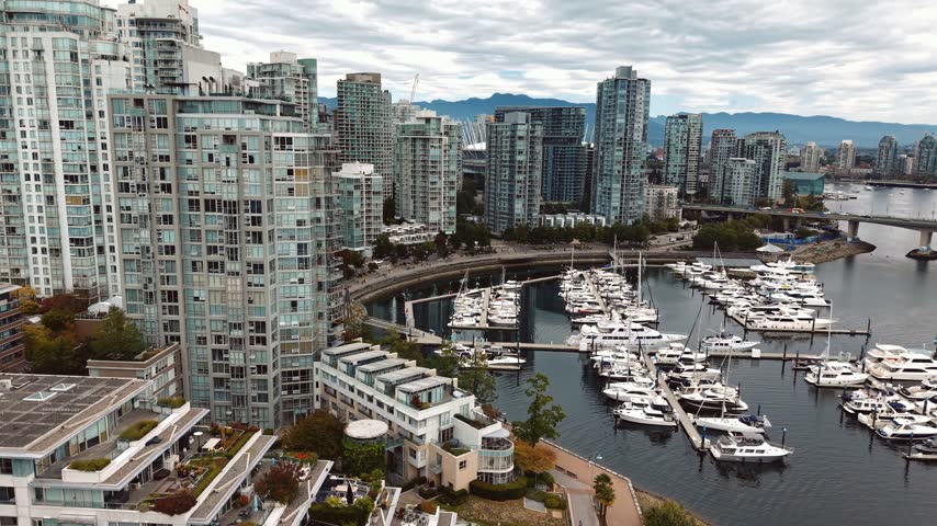 Aerial drone footage capturing a False Creek marina near the downtown stadium district, boats docked along a curved seawall path, condo towers and the city skyline beyond.