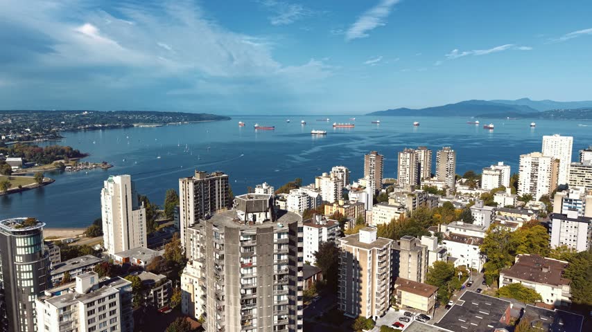 Aerial drone footage capturing English Bay on a sunny day, cargo ships at anchor and sailboats scattered across the water, West End towers and a sandy beach along the shoreline.