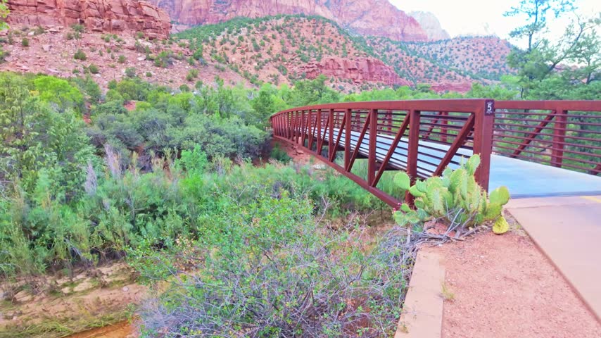 A bridge crosses the hiking trail with the colorful rock formations of Zion National Park rising behind