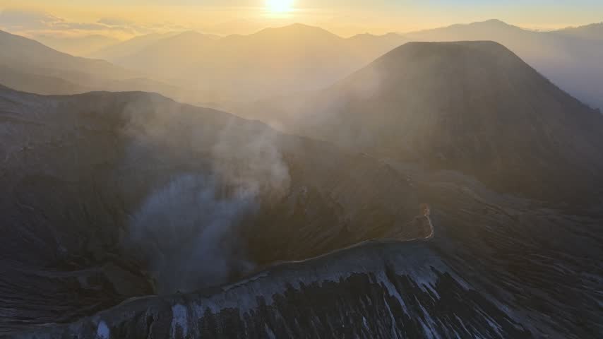 Golden sun beams over smoky, rugged volcanic Mount Bromo crater. Steam rises from crater