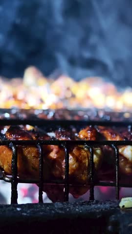 Vertical close-up of delicious meat and chicken wings grilling on a barbecue grate with rising smoke at night, handheld shot.