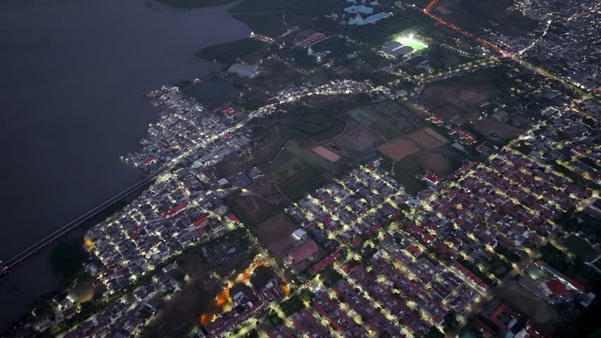 Dusk aerial drone view of Surabaya coastline and illuminated residential grid with river and stadium.