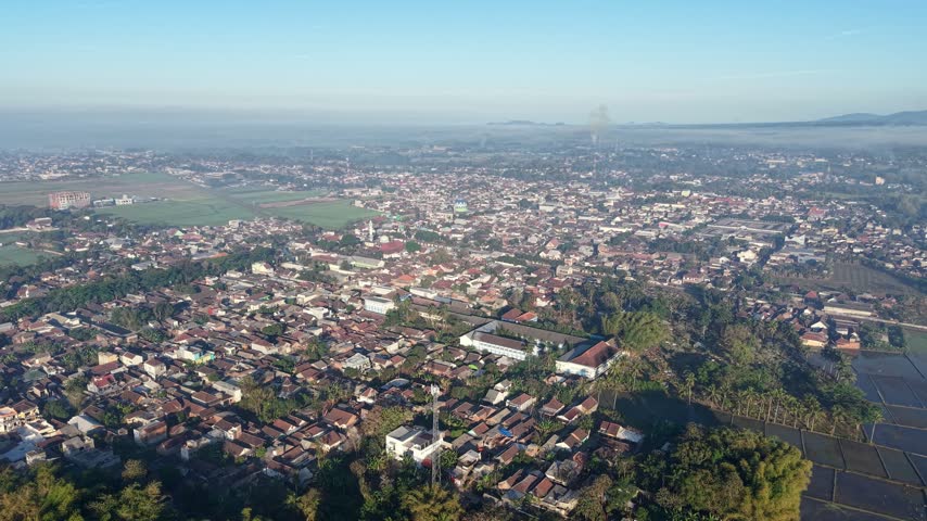 Expansive morning view of Kepanjen town with rooftops, rice paddies, misty hills and scattered greenery.