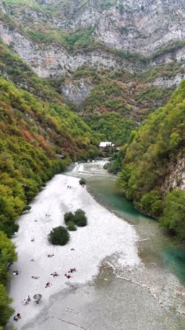 4K aerial drone footage looking upstream along the Shala River through the dramatic mountain valley, with people gathered along the riverbank and edge of the turquoise water.