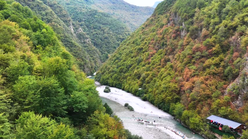4K aerial drone footage looking upstream along the Shala River through the dramatic mountain valley, with people gathered along the riverbank and edge of the turquoise water.