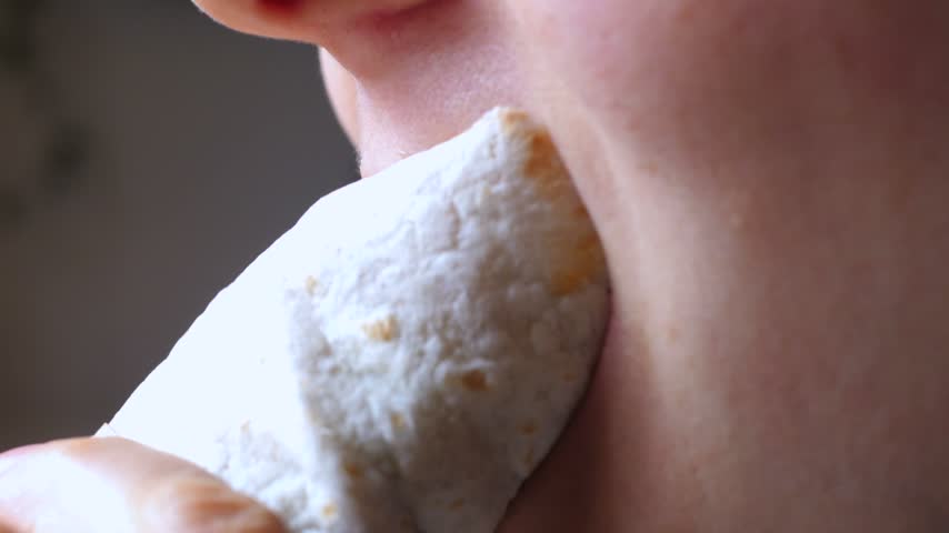 Person bites wrap near mouth, holding stuffed flatbread close while chewing. Close up food moment emphasizes lips, skin texture, and tender movement with quiet, satisfied mood in warm light.