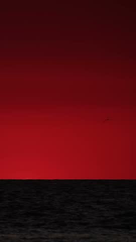 The sun sets over the ocean, creating vibrant shades of orange and red in the sky reflecting on the water. The horizon showcases a transition of light as the sun descends.
