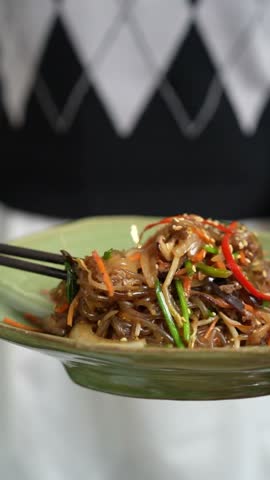 Close up of chopsticks lifting Korean japchae glass noodles with vegetables and beef from bowl.