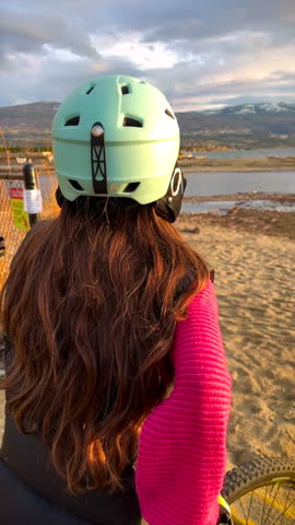 Vertical format of a young girl wearing a helmet, taking a moment looking around at the scenery in front of her, with sand, water, mountain and an overcast sky in the background