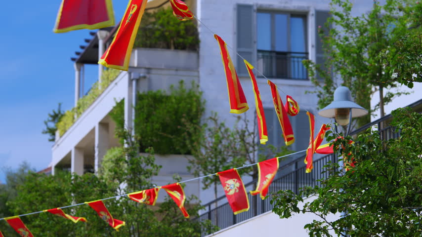 A string of red and yellow flags of Montenegro flutters in the wind against a white multistory building with balconies and green trees, decorating an outdoor space for a celebration or diplomatic.