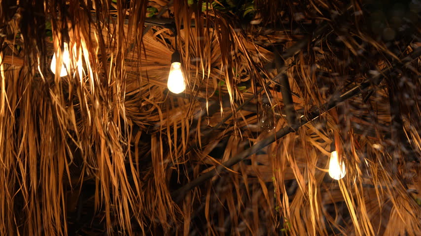 Warm bulb string lights hang beneath a straw thatched roof, swaying and flickering as strong wind and an approaching storm shake the dried palm leaves at night.