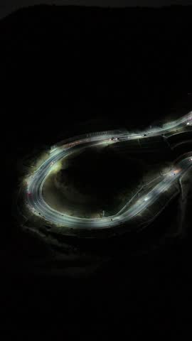 Aerial night view of a winding mountain road illuminated by streetlights and vehicle headlights, with a sprawling city glowing across the valley under a dark sky.