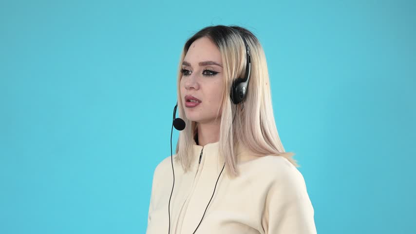 A young blonde woman wearing a headset with microphone speaks while working as a call center operator against a bright blue background.