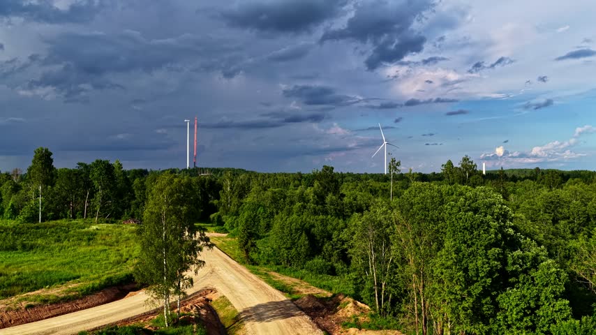 Wind turbines generating renewable energy in a green forest with a dirt road under a stormy sky