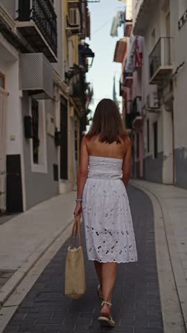 A person walks alone down a narrow street lined with buildings. The sun is shining and the atmosphere is lively. The street features shops and cafes.