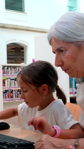 Elderly grandmother lovingly teaching her young granddaughter how to use a computer, sharing knowledge and creating a strong family bond between different generations inside a public library