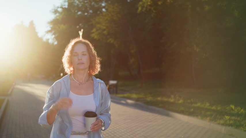 A woman with visible dental braces walks through a park with takeaway coffee, adjusts her hair, and smiles in evening light.