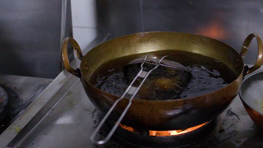 Oil bubbles vigorously inside a brass wok on a gas flame in a commercial kitchen.