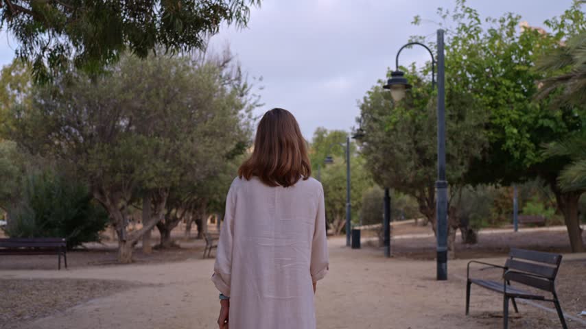 A person walks along a pathway in a park during dusk. The area features trees and benches on either side. The sky shows some clouds as daylight fades.