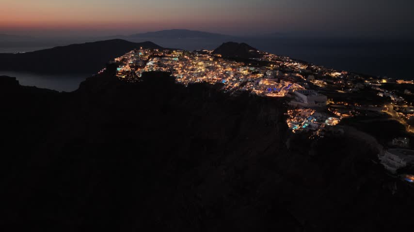 Sparkling lights illuminate the town of Fira on the island of Santorini in Greece during the quiet blue hour of a peaceful evening.