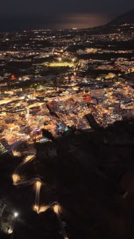 Bright lights shine across the town of Fira in Santorini Greece as evening darkness falls over the Mediterranean landscape with glowing pathways
