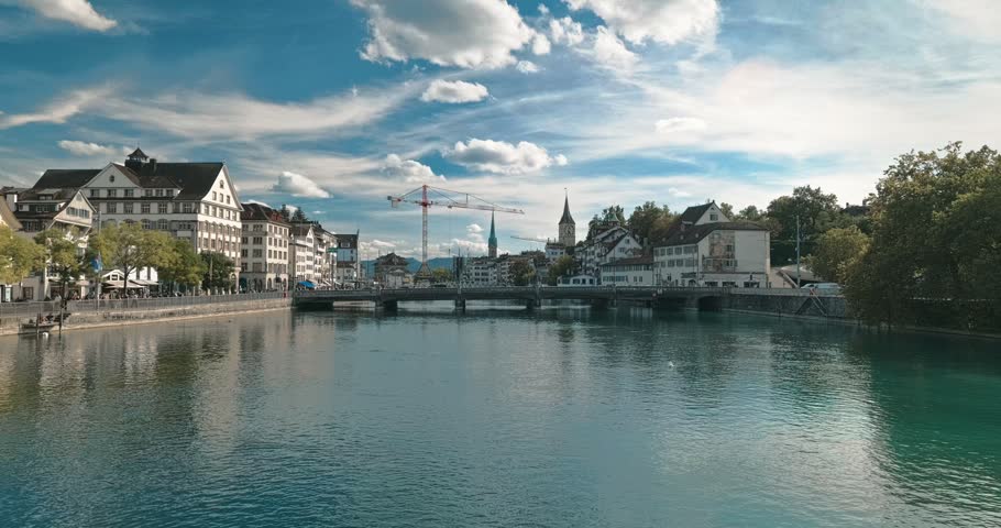 Wide static shot of the Limmat River in Zurich, Switzerland, featuring historic architecture, bridges, and a working construction crane moving in the background under a blue sky with fluffy clouds.