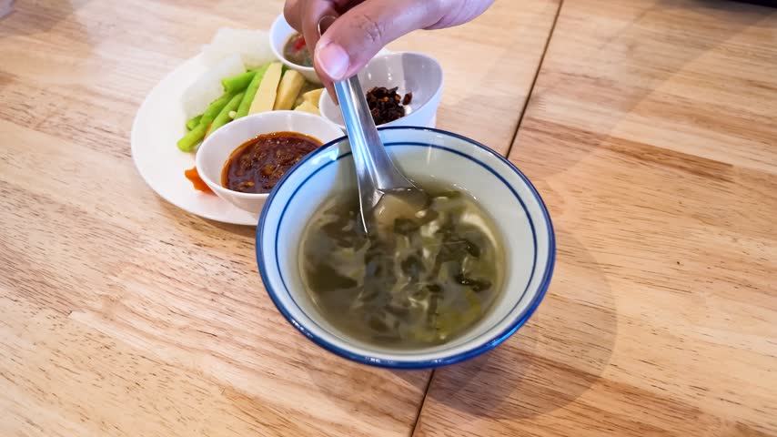 Top-down view of a hand stirring hot traditional Burmese soup on a wooden table.