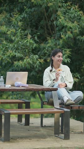 Young woman sitting cross-legged at a picnic table in a park, enjoying a donut during a break from working on her laptop outdoors