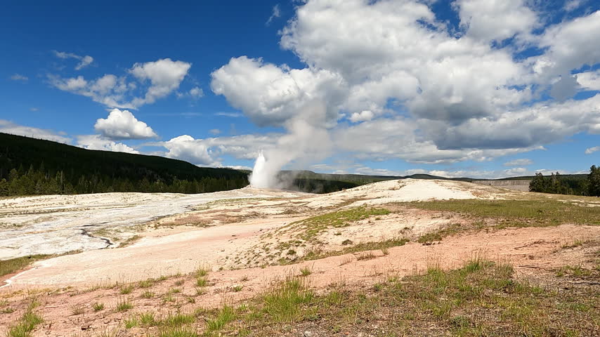 Old Faithful geyser erupting a tall column of steam and water under a cloudy blue sky.