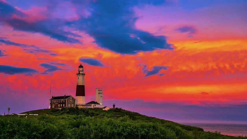 An elevated view captures the historic Montauk Point Lighthouse set against a vibrant sunset sky on the eastern tip of Long Island, New York. The composition surveys coastal architecture, beacon structures, surrounding vegetation on the headland, and dramatic red and blue twilight cloud formations under evening conditions. This travel, architectural, and regional landscape asset suits historical documentaries, American tourism promotions, maritime presentations, and cinematic establishing sequences requiring East Coast geography.   