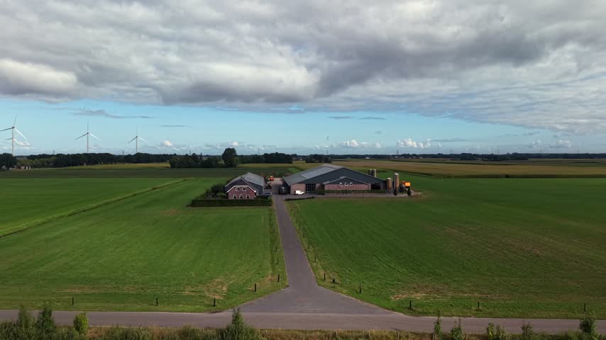 Brick farmhouse connects to industrial barn with grain silos, straight paved driveway cuts through green fields, wind turbines rise beneath dramatic clouds in Netherlands. Aerial drone wide shot