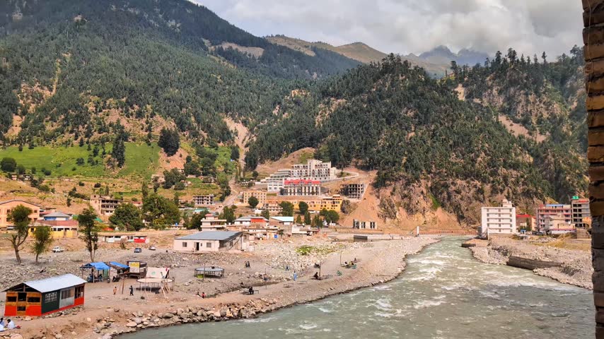 Mountain river landscape with small town buildings and green hills under a cloudy sky
