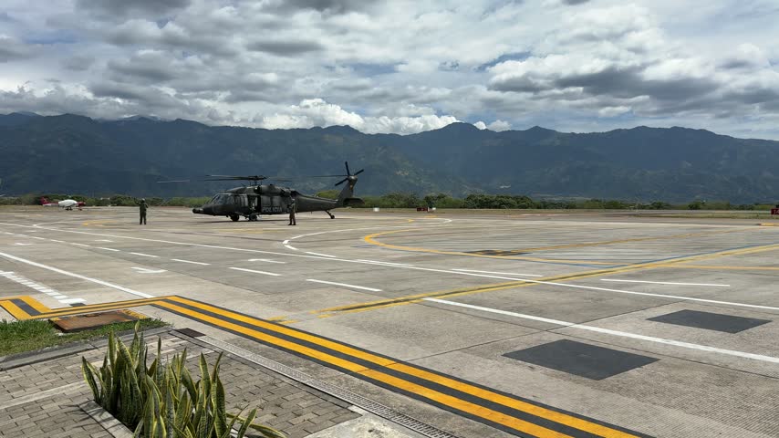 A black Colombian National Police helicopter taxis across the apron, followed by a bright green airport fire rescue truck driving on the tarmac at Perales Airport in Ibagué, Colombia, backed by distan