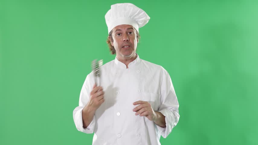 Adult male chef in white cook uniform and hat stands against green screen with pastry tongs and explaining dessert preparation or baking method 