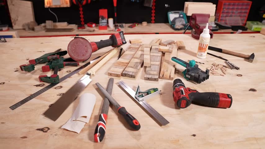 A collection of woodworking tools including a cordless drill, sander, hand saw, clamps and measuring tools lies beside timber pieces on a carpentry workbench.