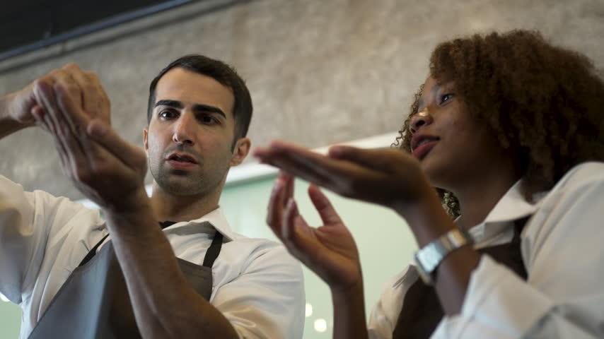 Education in coffee tasting: a man sniffs his cupped hand while a smiling teenage girl offers her open palm, sharing aroma learning in a barista training class in a cafe.