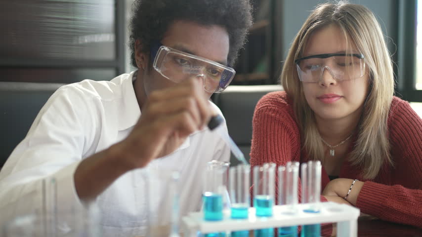 A young man uses a pipette over test tubes with blue liquid while a smiling teenage girl watches, both in safety goggles. Education, science class and teamwork concept with a positive mood.