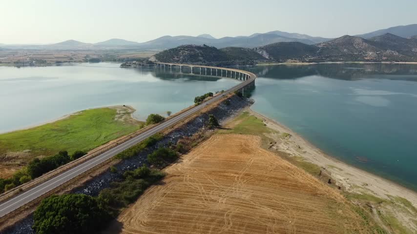 Breathtaking cinematic drone shot capturing the iconic long concrete bridge and winding road crossing the beautiful blue water reservoir of Polyfytos Lake in Kozani.