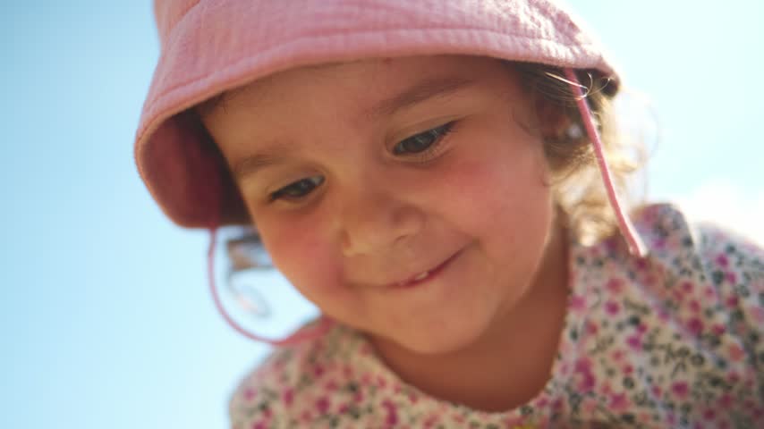 Young girl in pink hat smiles while holding tiny flower. Soft sunlight highlights her joyful expression. Focus on her hands delicately handling the bloom. Ideal for nature, childhood