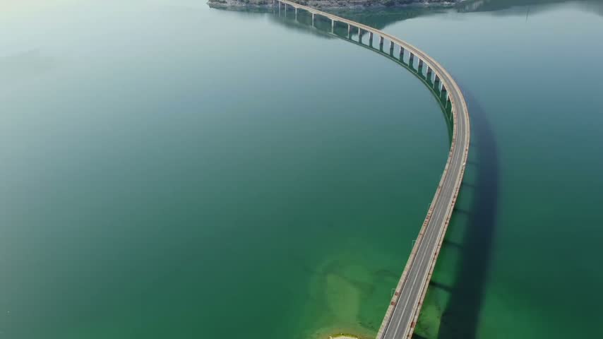 High altitude cinematic drone shot capturing the perfect architectural S-curve design of the long concrete bridge crossing Polyfytos Lake reservoir in Kozani.