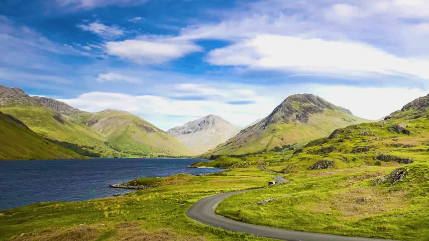 Moving clouds drift over rugged green fells and a tranquil body of water.