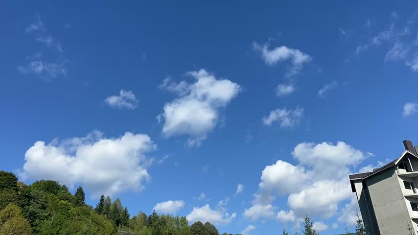 Timelapse of white clouds moving across a bright blue sky above treetops and a mountain area.